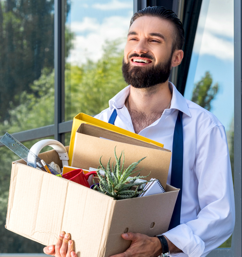 A bearded, male high performer with his tie untied, walking out the door after quitting, and carrying a box of his things