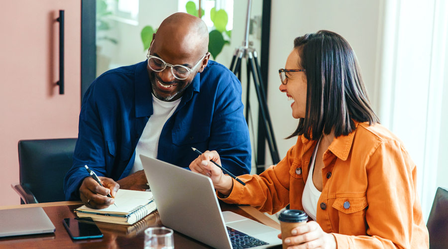 Two hiring managers, a Black man writing notes and an Asian woman sitting in front of a laptop, discussing the environment fit scores for the candidate's they are about to interview