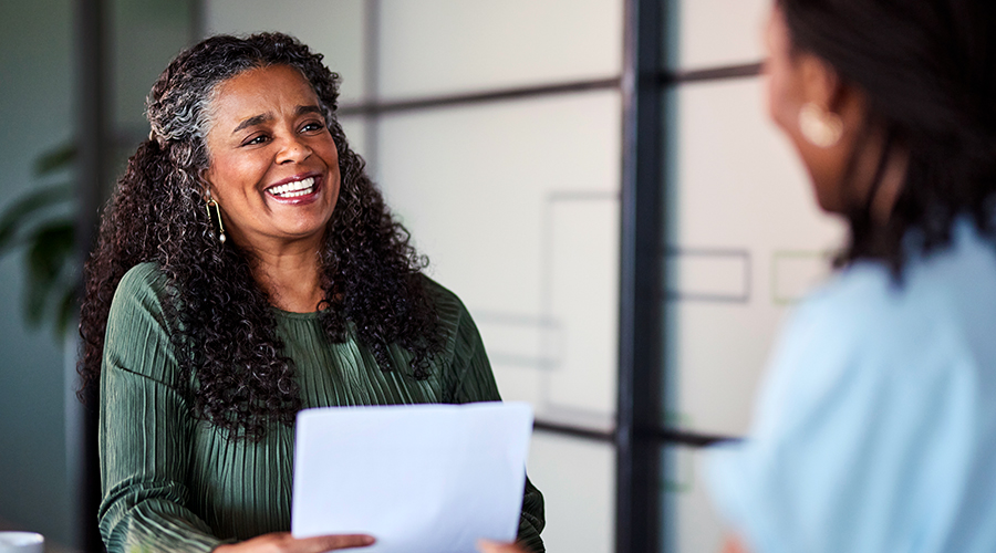 A Black, female, senior -level manager interviewing a Black, female candidate for an open position, and smiling because the manager and candidate are compatible